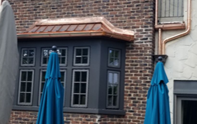 Bay window with copper roof and dark gray frame on brick building. Two blue umbrellas in foreground.