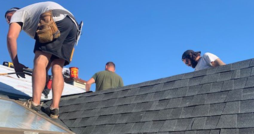 Three workers on a rooftop, installing shingles on a sunny day.