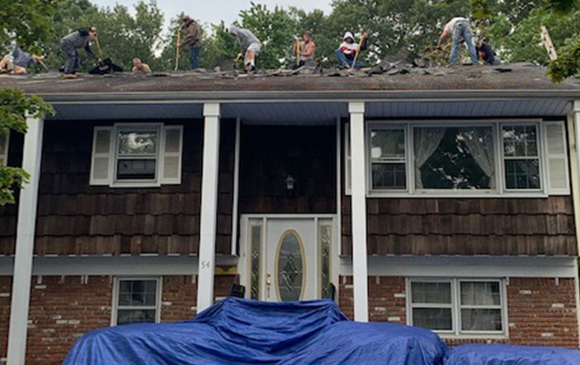 People on a roof removing shingles, with a car covered by a tarp in front of a house.