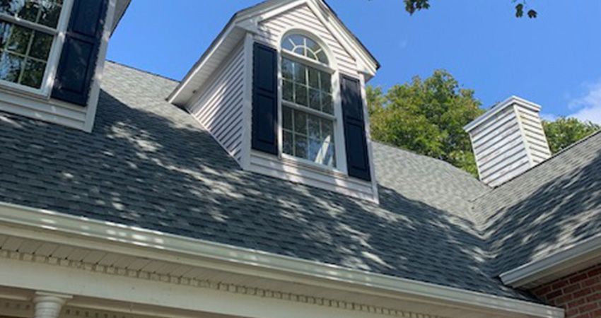 A gray-shingled roof with a dormer and chimney, seen against a clear blue sky.