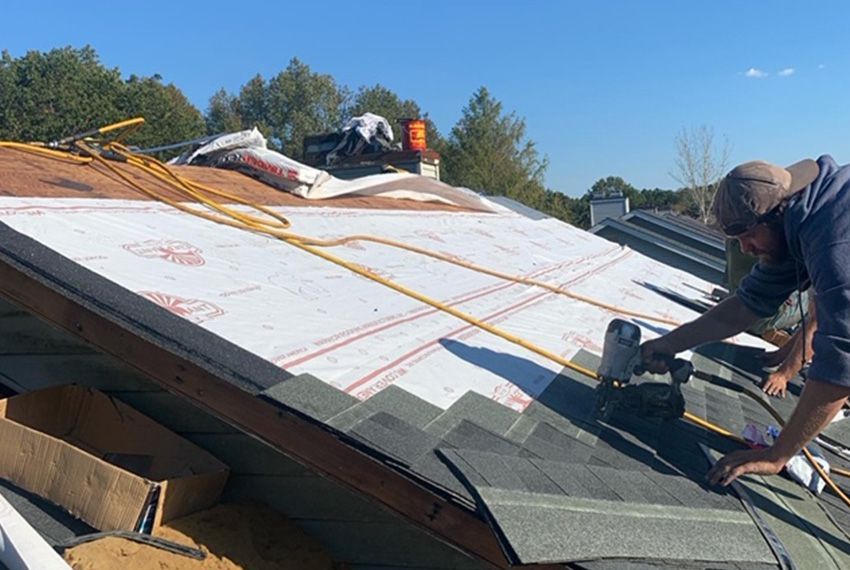Person nailing shingles onto a roof, with protective underlayment visible. Sunny outdoors.