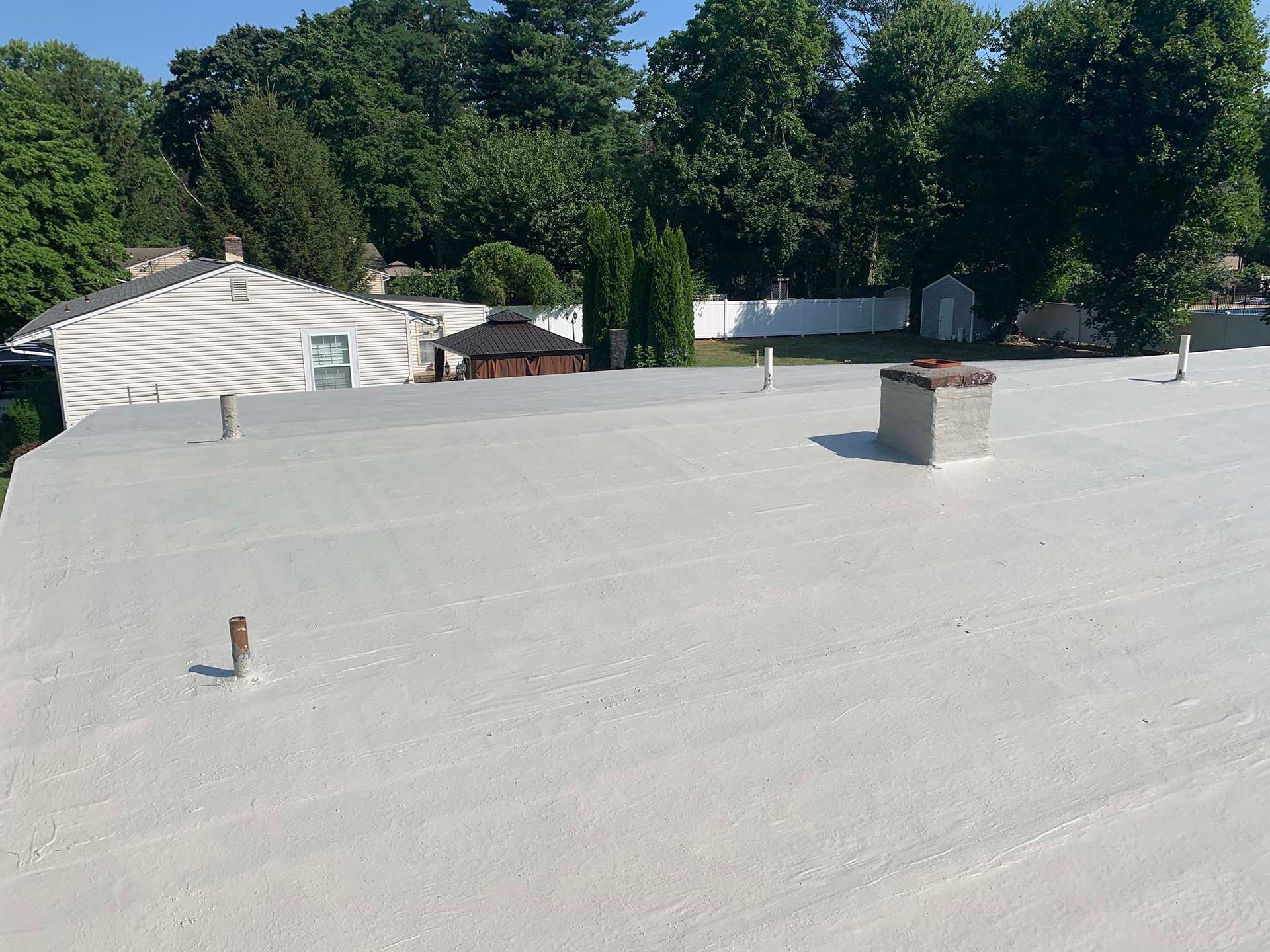 White flat roof with chimneys and vents, trees and houses in the background on a sunny day.