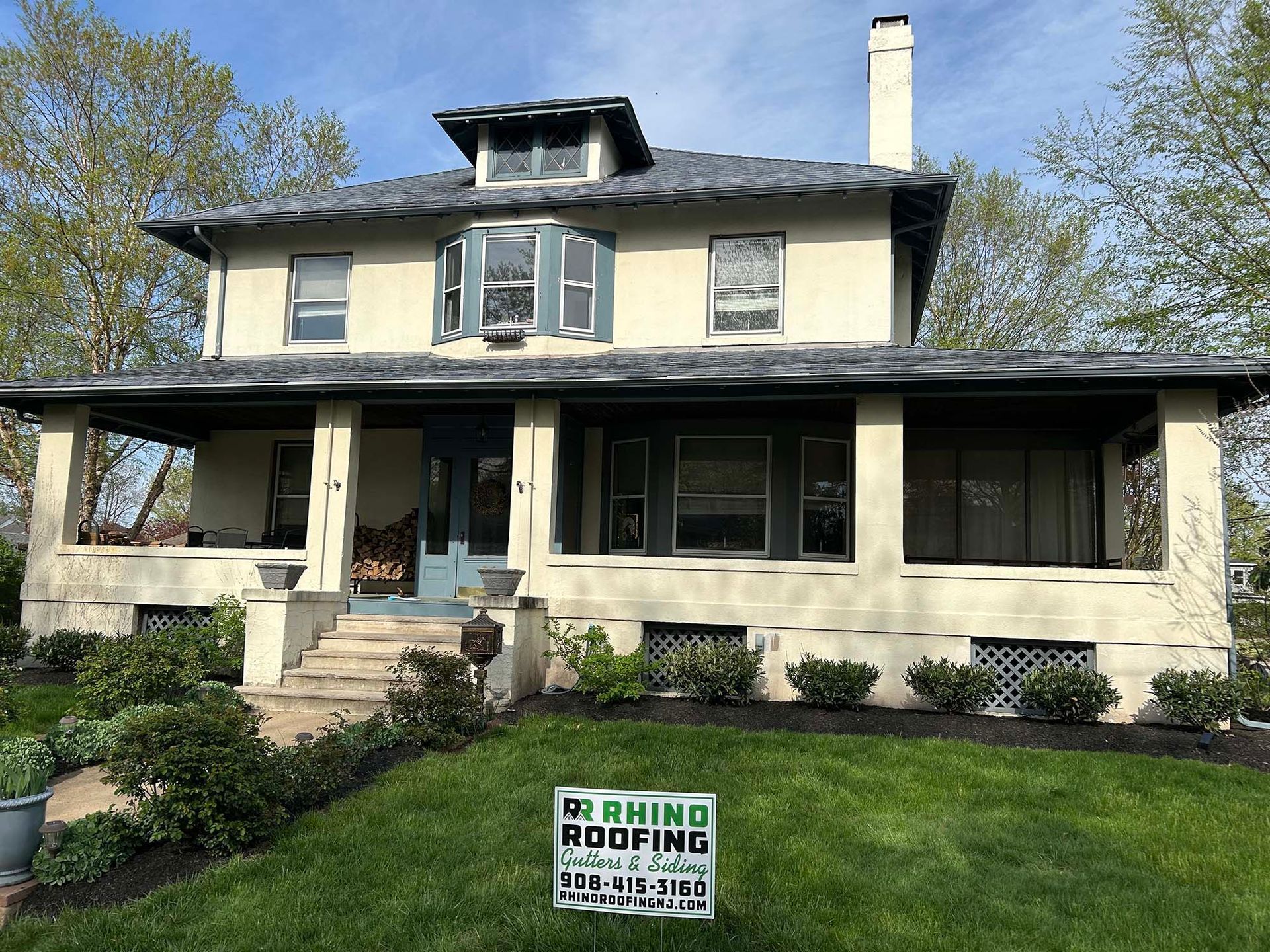 Two-story beige house with a dark gray roof. A wraparound porch and front lawn are in view.