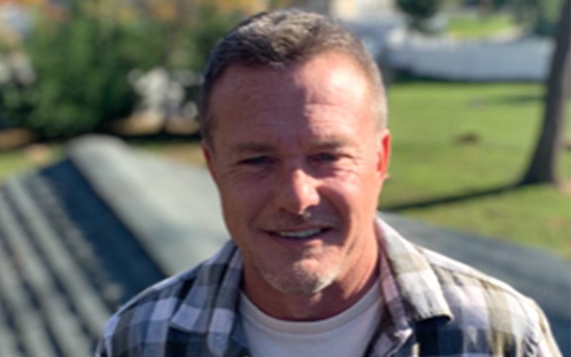 Man smiling outdoors, wearing a plaid shirt over a white t-shirt. Green background with roof tiles in front.