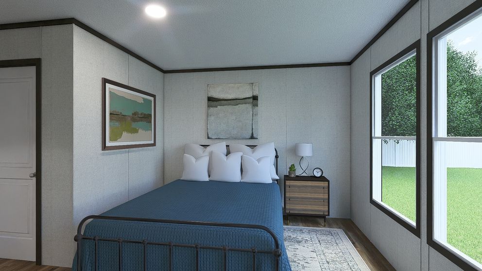 Modern bedroom with a blue bedspread, bedside table, framed art, and large windows overlooking a green yard