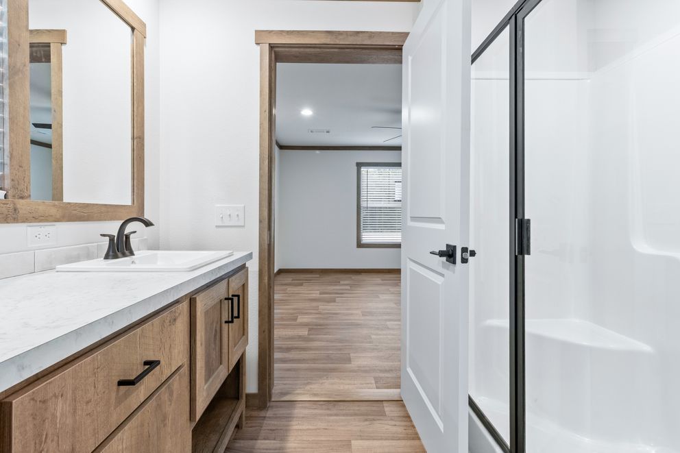 Modern bathroom with wood vanity, white countertop, mirror, and open doorway to a hallway or adjoining room