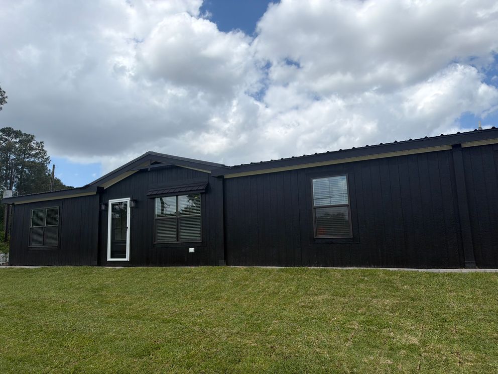Black single-story building with white-trimmed windows and door, set behind a grassy lawn under a cloudy sky