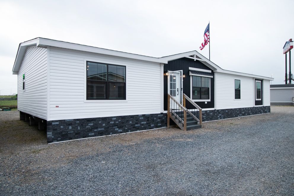 White modular building with black stone base, front porch, and an American flag on gravel lot