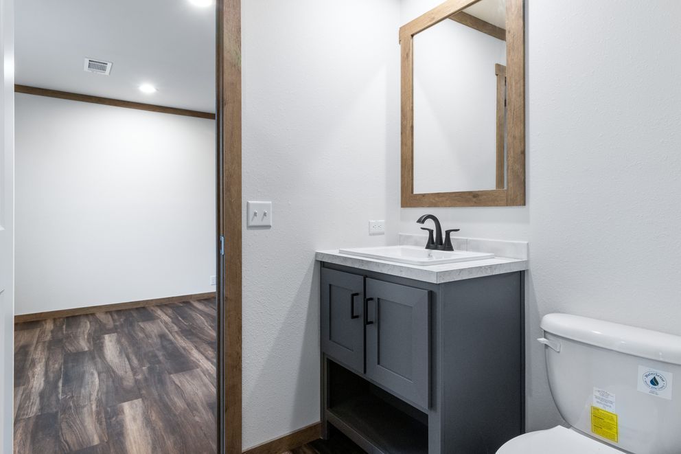 Modern bathroom vanity with gray cabinet, white sink, toilet, and wood-framed mirror beside an open doorway.
