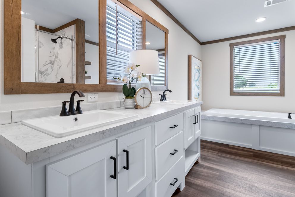 Bright bathroom with double vanity, black fixtures, wood-framed mirrors, and a bathtub by the window