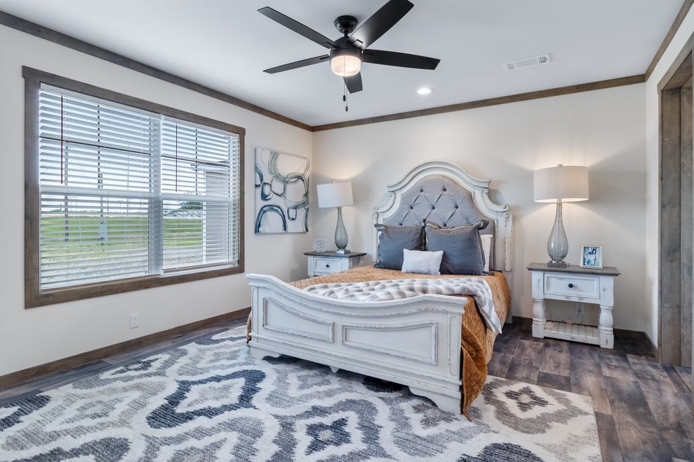 Bright bedroom with a white ornate bed, gray-blue rug, ceiling fan, and window blinds.