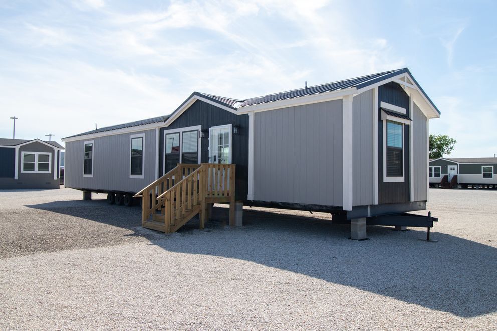 Gray mobile home with wooden steps on a gravel lot under a bright sky