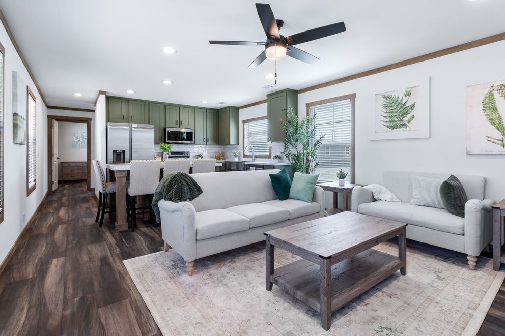 Bright open-concept living room with gray sofas, coffee table, dining area, and kitchen in the background.