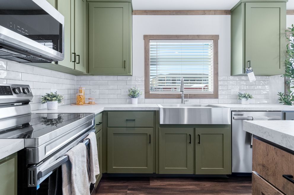 Bright kitchen with green cabinets, white tile backsplash, stainless stove, and window over the sink.
