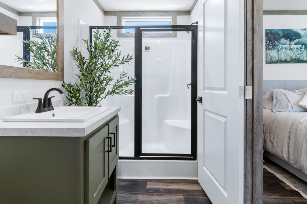 Modern bathroom with glass shower, green vanity, and bedroom visible through open door