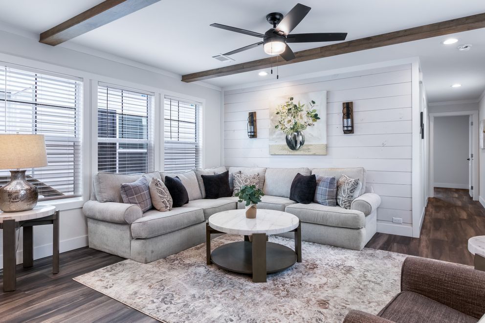 Bright living room with gray sofas, round coffee table, ceiling fan, and large windows with blinds.