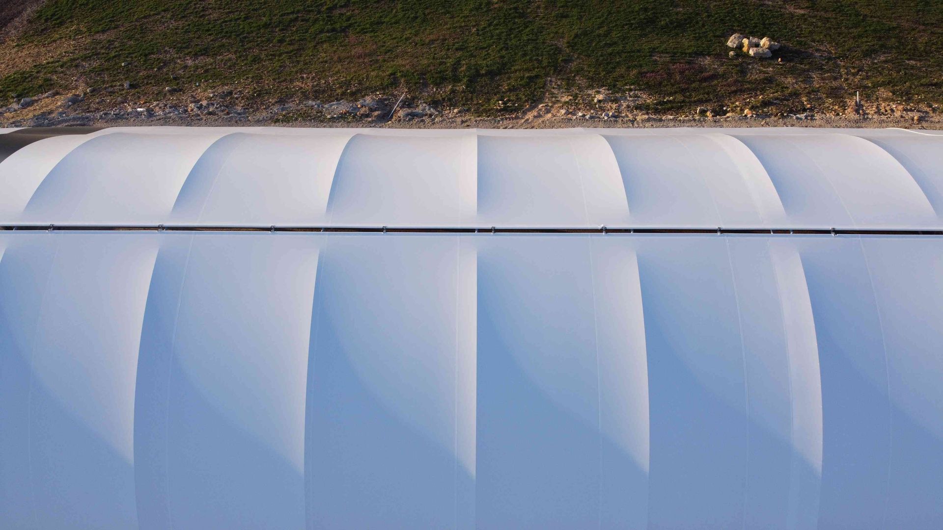 White, arched structure with segmented panels, viewed from above, with green hillside in the background.