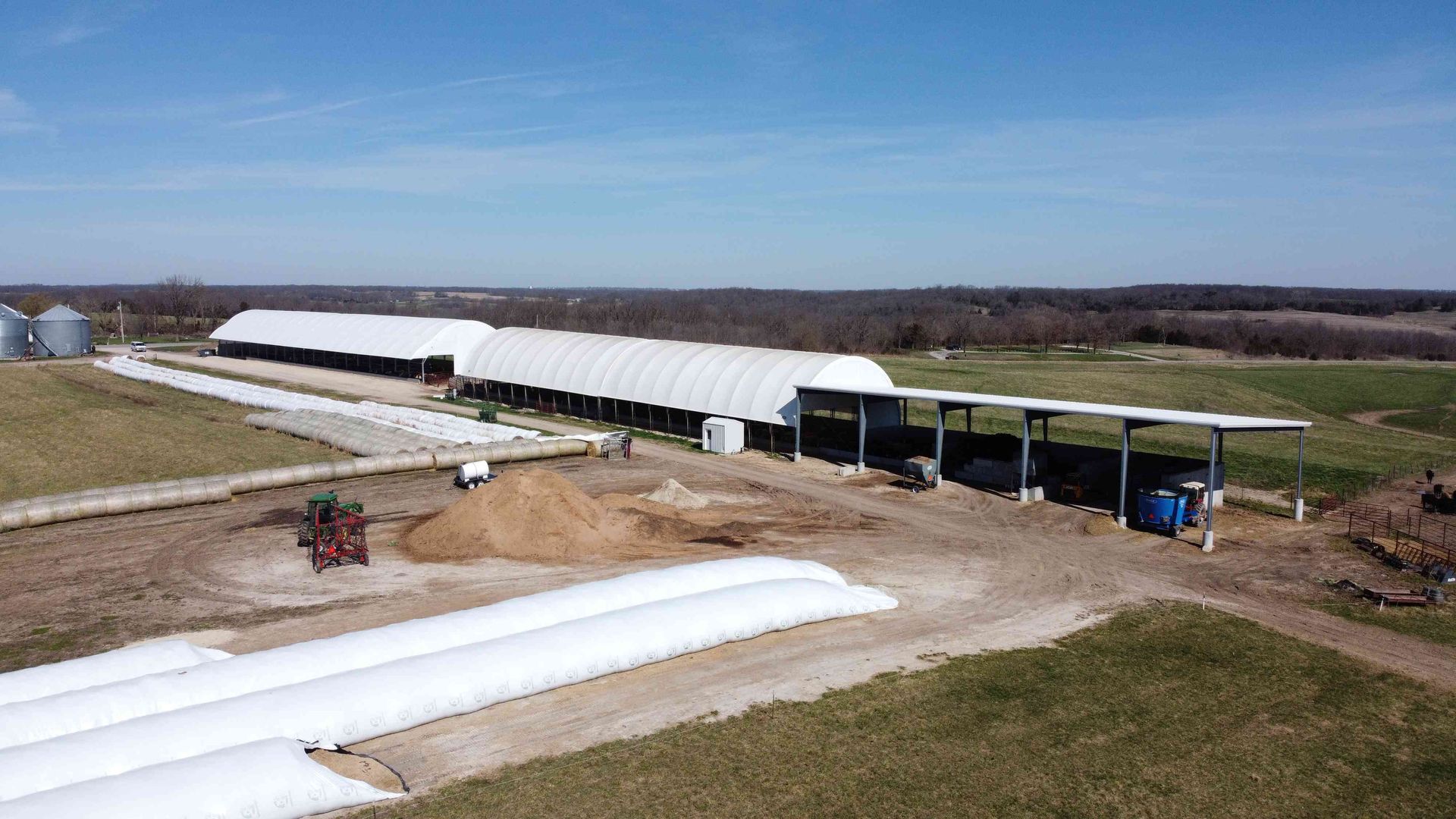 Farm buildings with white arched roofs and silage bags on a grassy field under a blue sky.