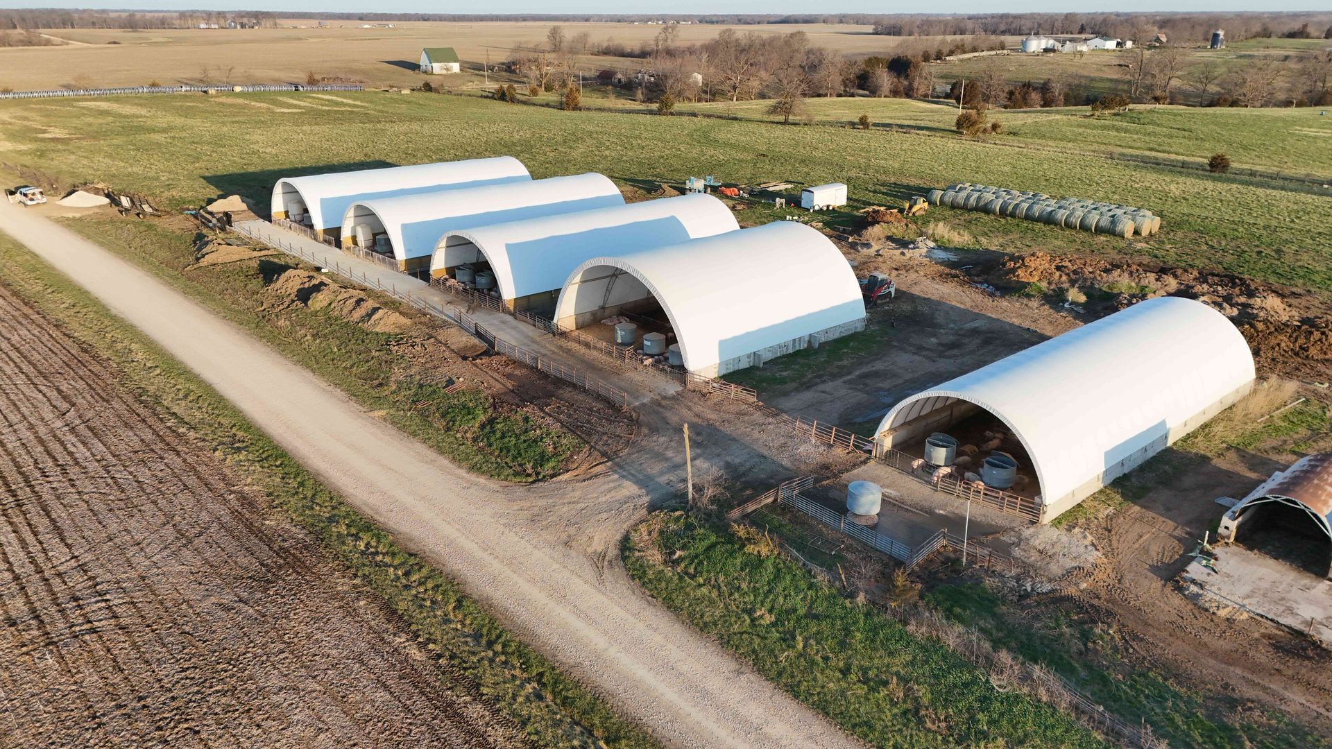 Row of white arched-roof farm buildings beside a dirt road in a field.