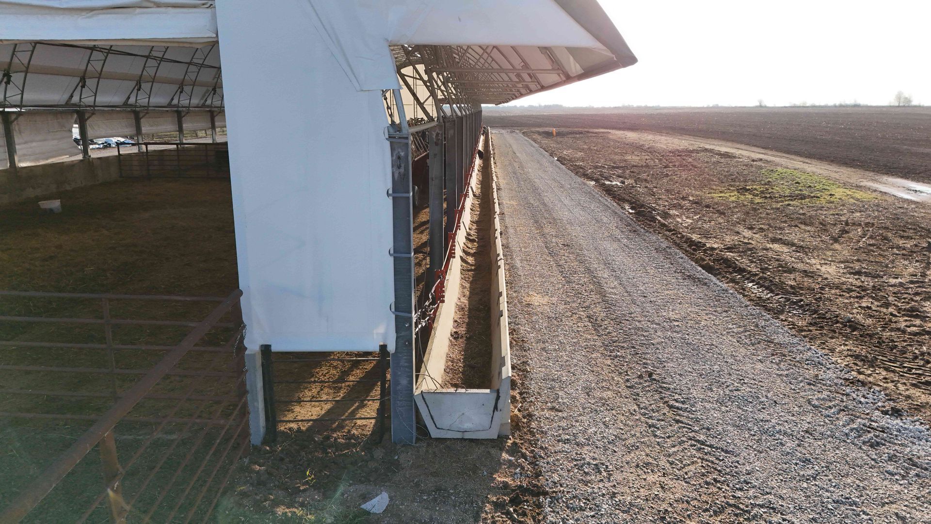 Barn with concrete feeding trough, gravel path, and open field under a sunny sky.