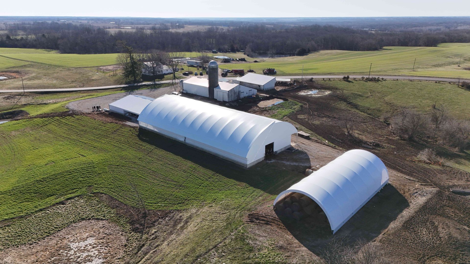 Aerial view of a farm with a large white arched building and silo, in a green field.