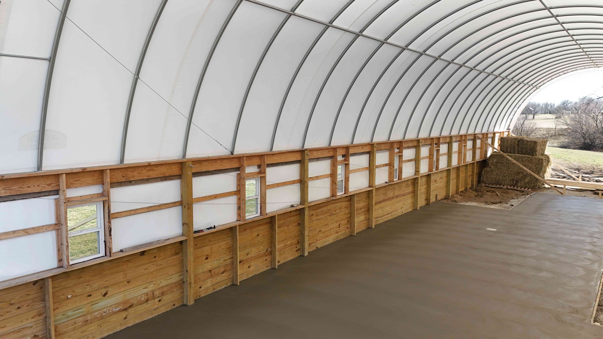 Inside view of a large, unfinished building with a concrete floor, wooden walls, and a curved, white ceiling.