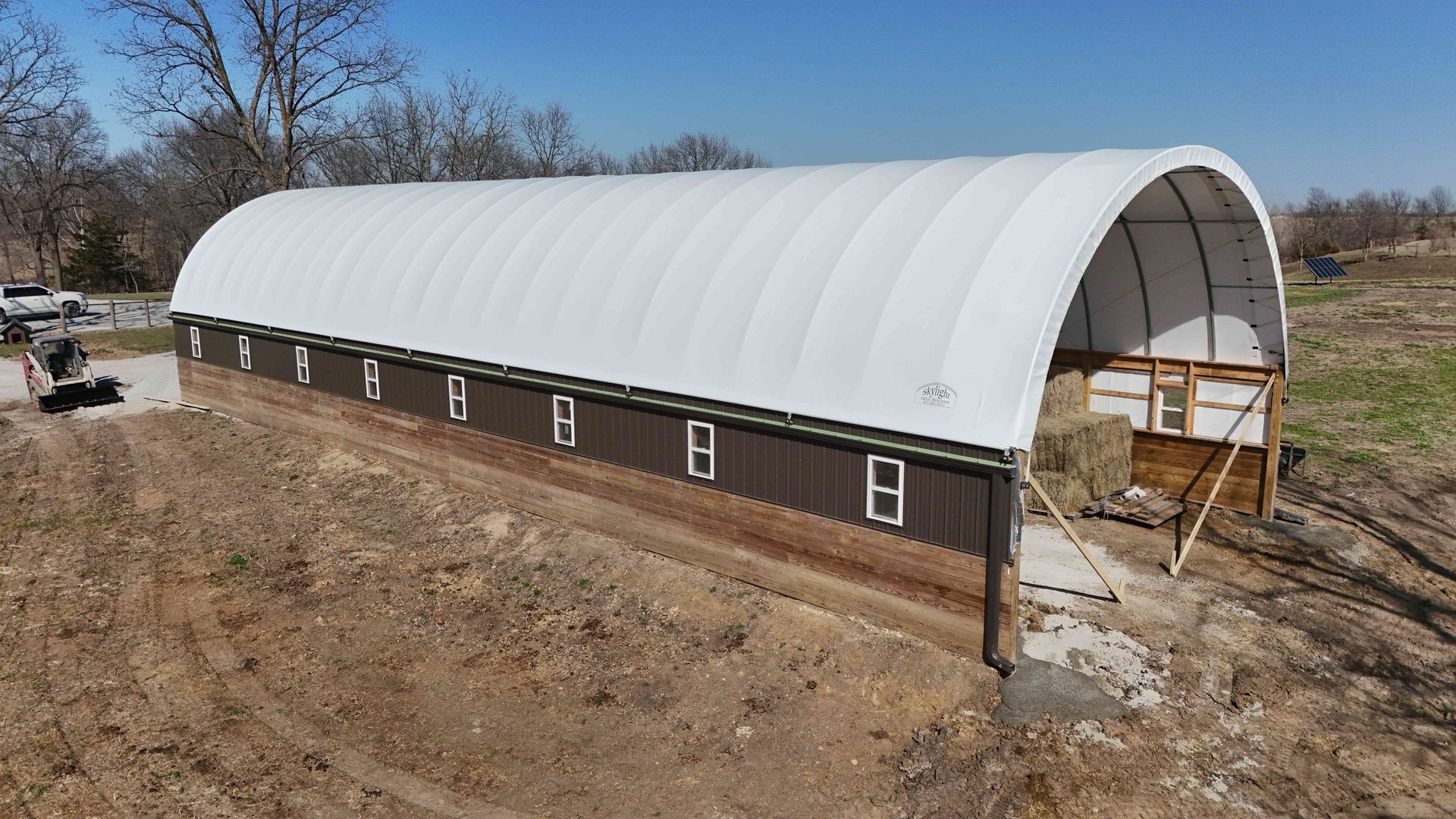 White arched roof over a brown-sided barn on a dirt lot.