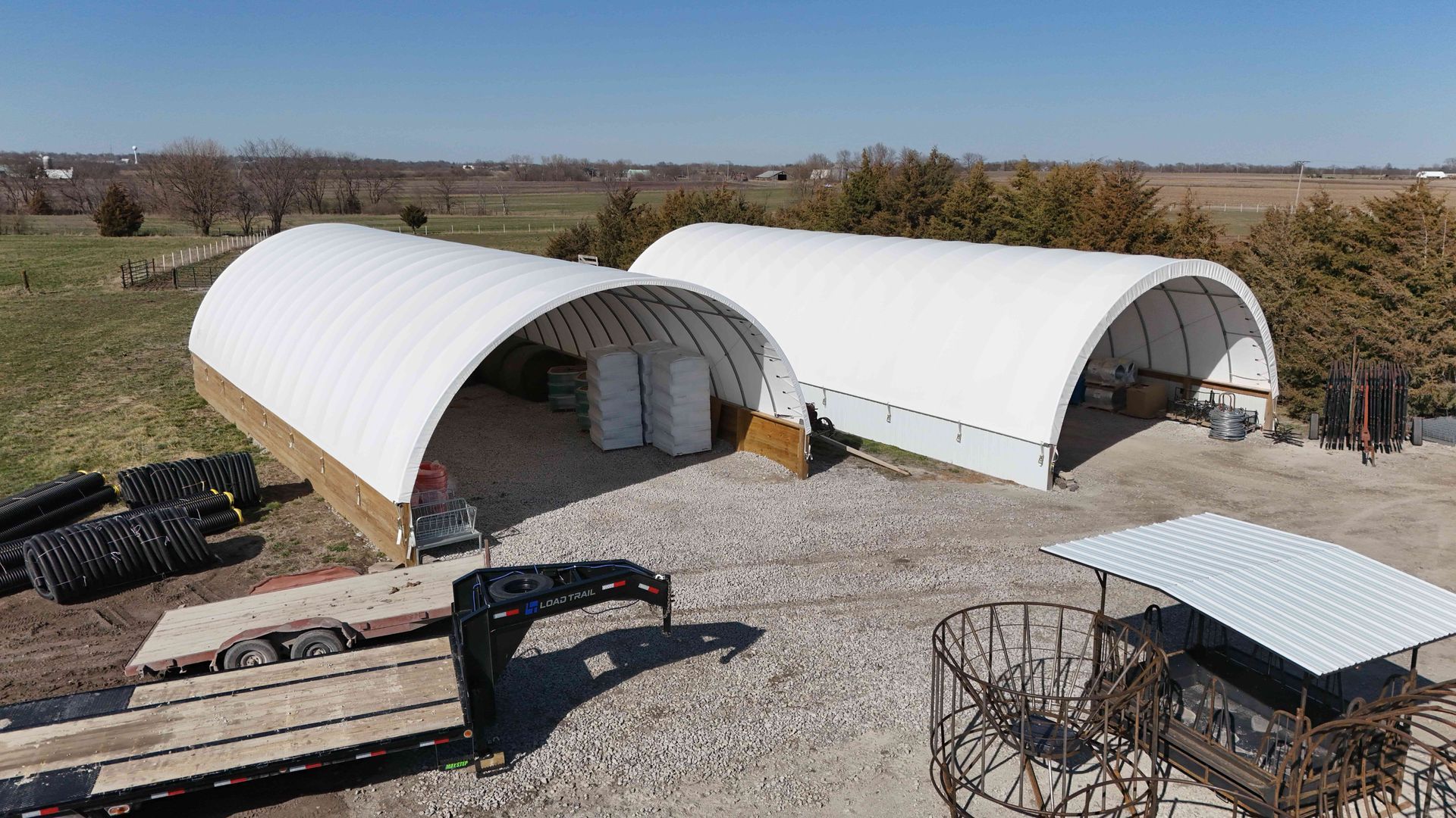 Two white arched-roof storage structures on a gravel surface, with farm equipment, trees, and blue sky.