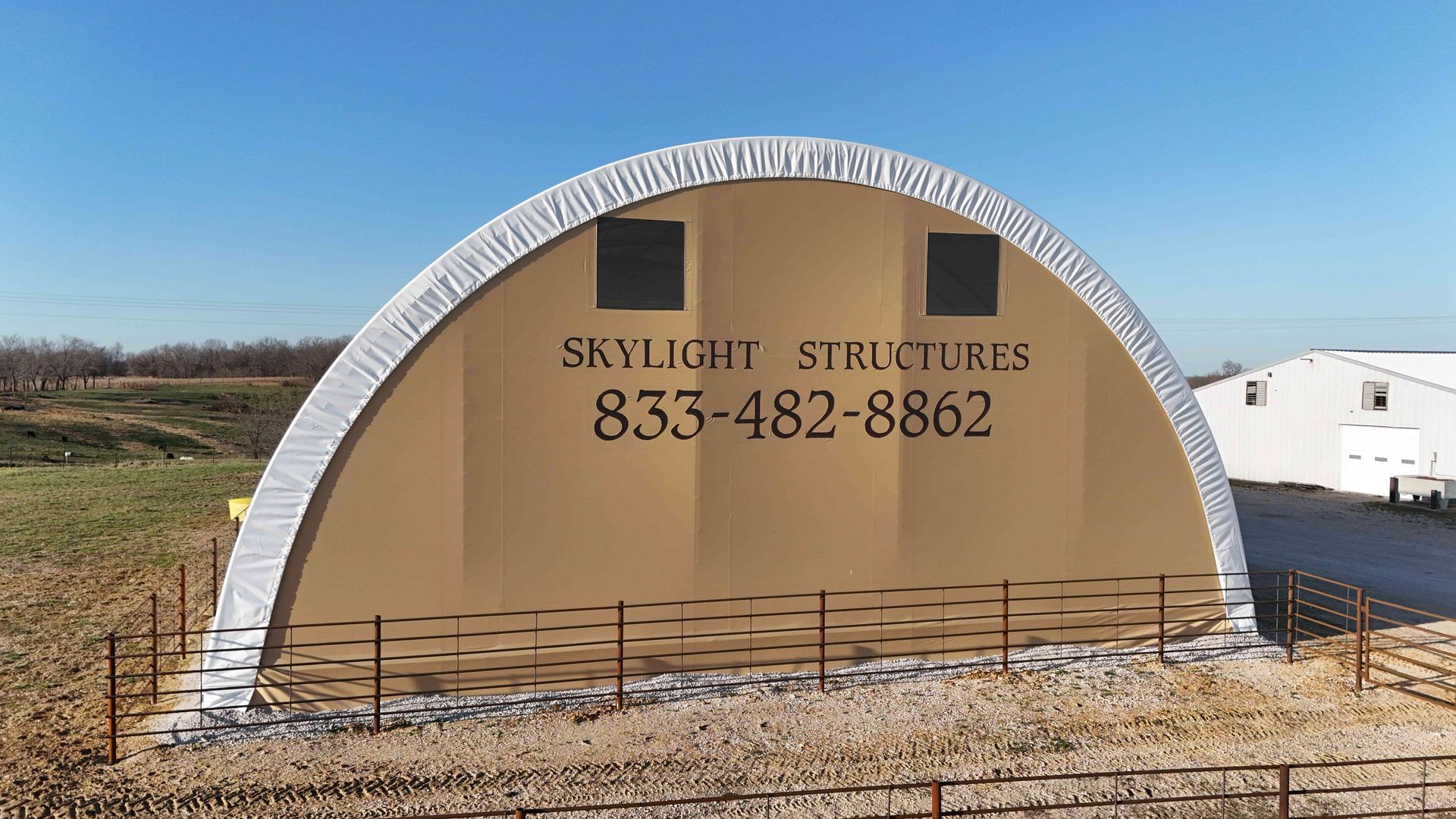 Tan arched structure with Skylight Structures logo and phone number. Two square windows, set in a field.