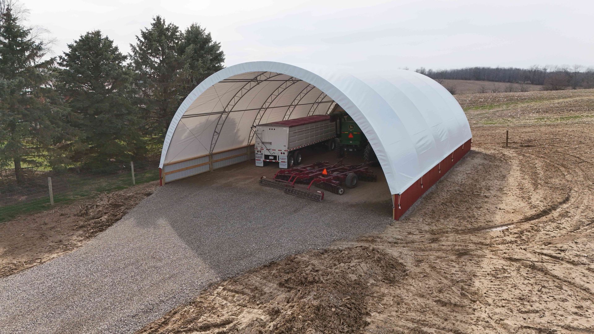 White arched-roof structure on a gravel base in a field; red trim and trees in the background.