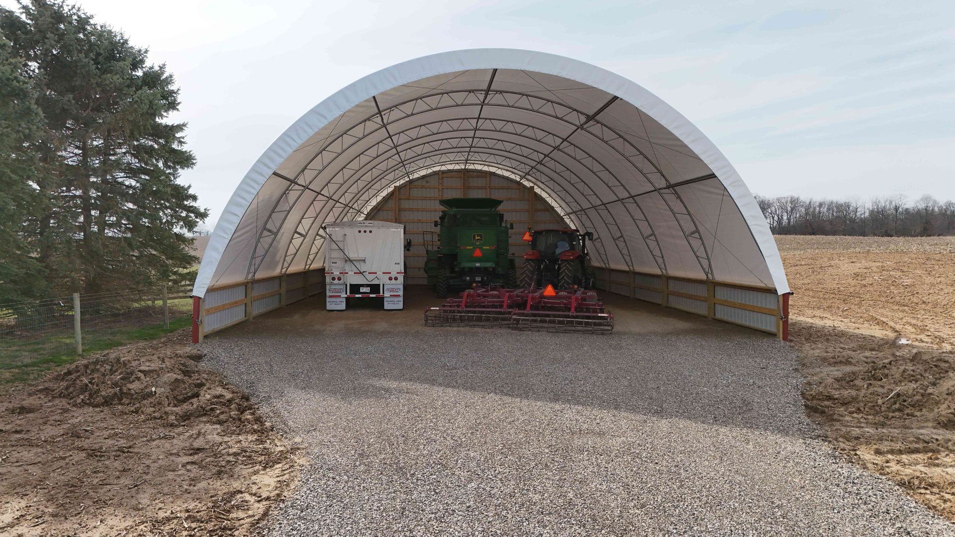 White arched farm building with tractor, trailer, and implements on gravel.