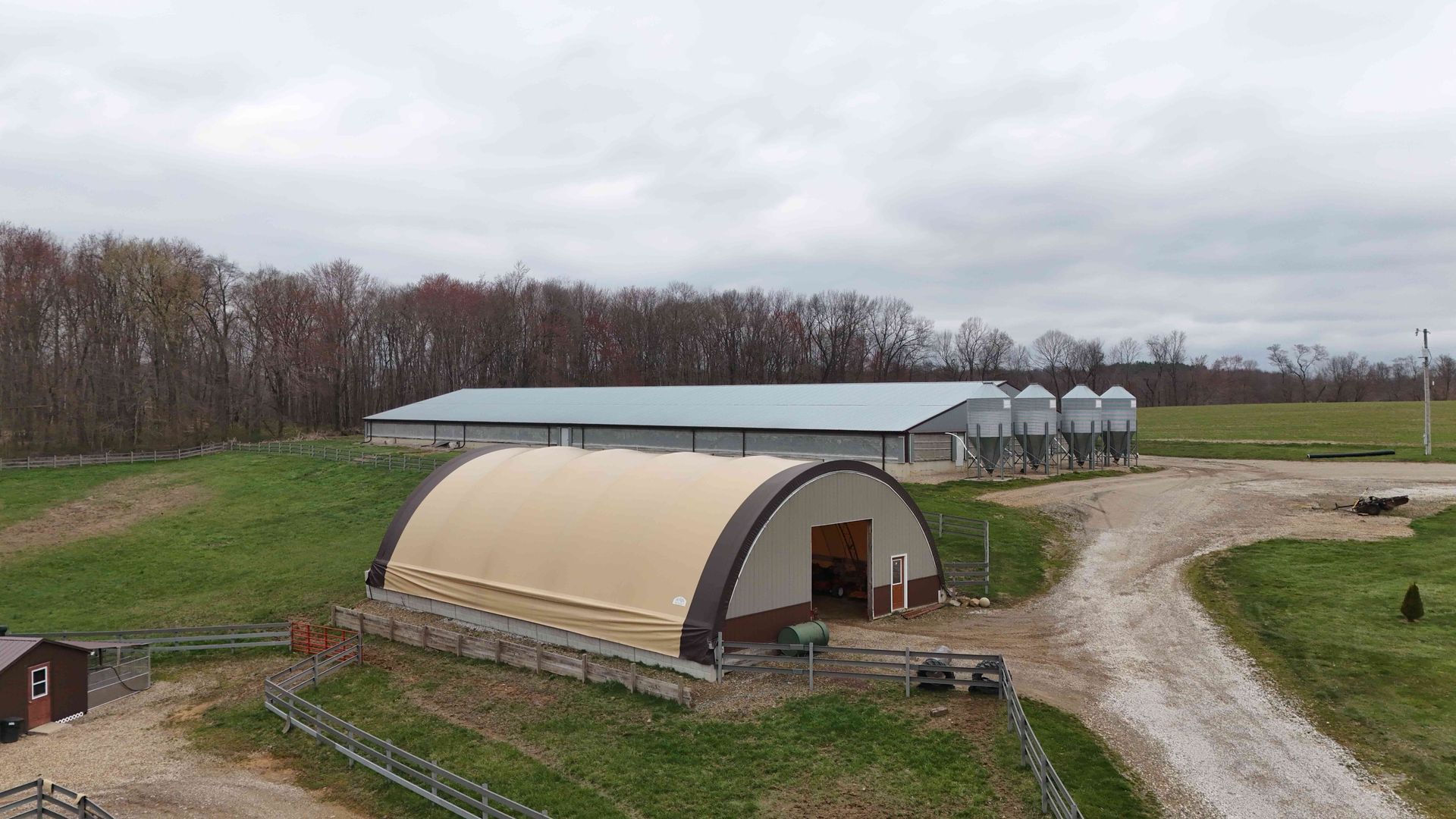 Farm buildings on a green hillside under a cloudy sky. A large arched shed is in the foreground, and other buildings are behind it.