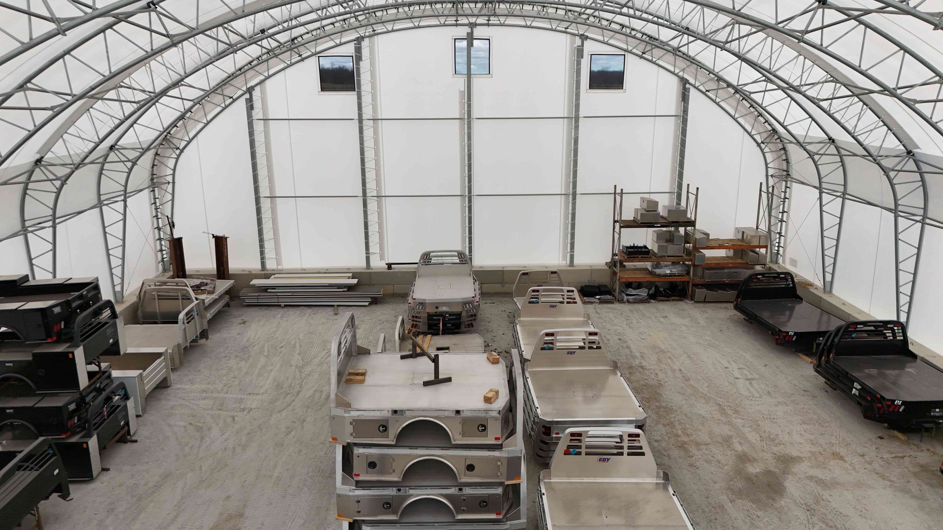 Inside a white arched industrial building, several truck beds are in a row, with a workshop area in the background.
