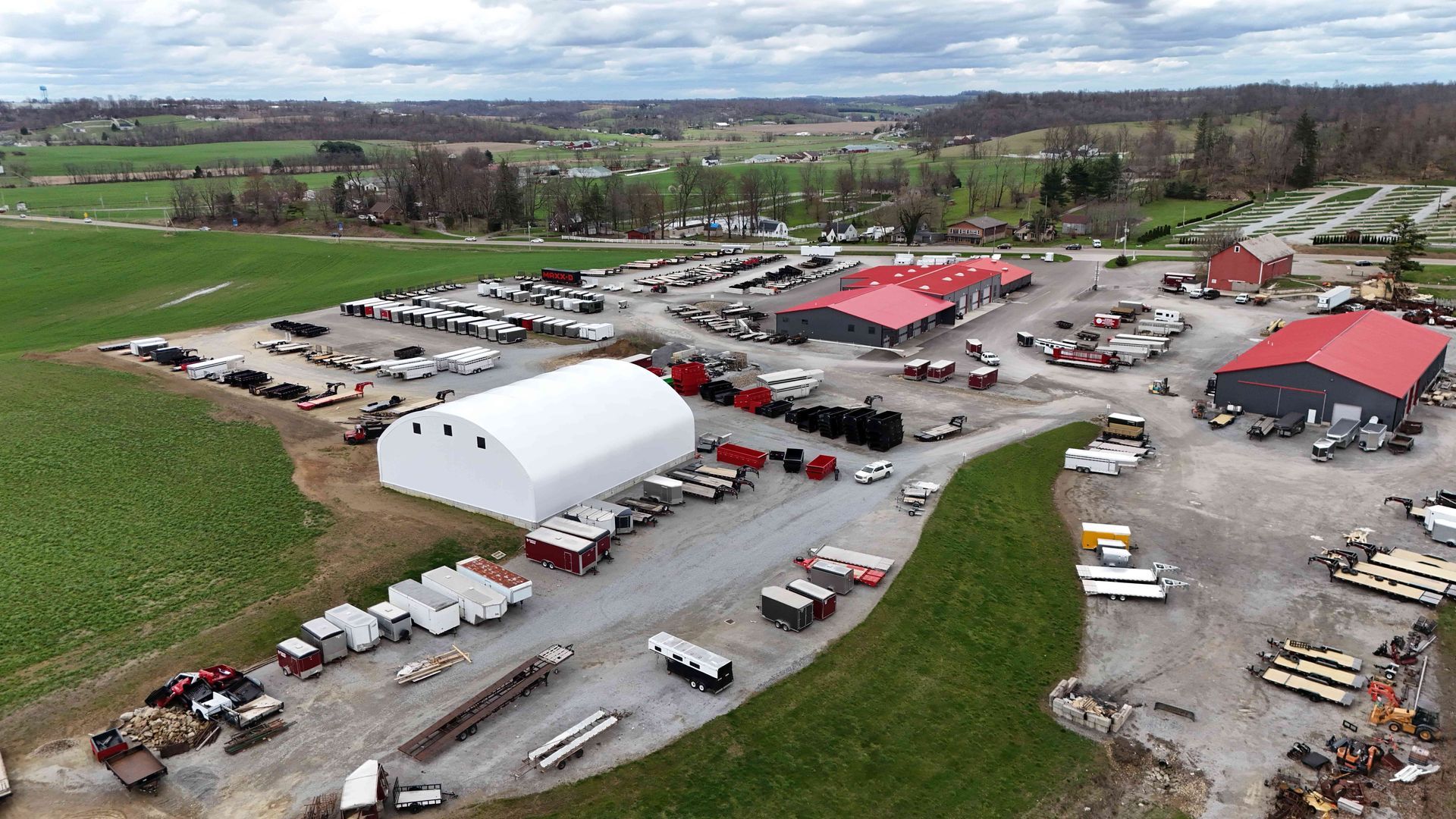 Aerial view of a business complex. Structures with red and black roofs, white tent, vehicles parked outside, green fields surrounding.
