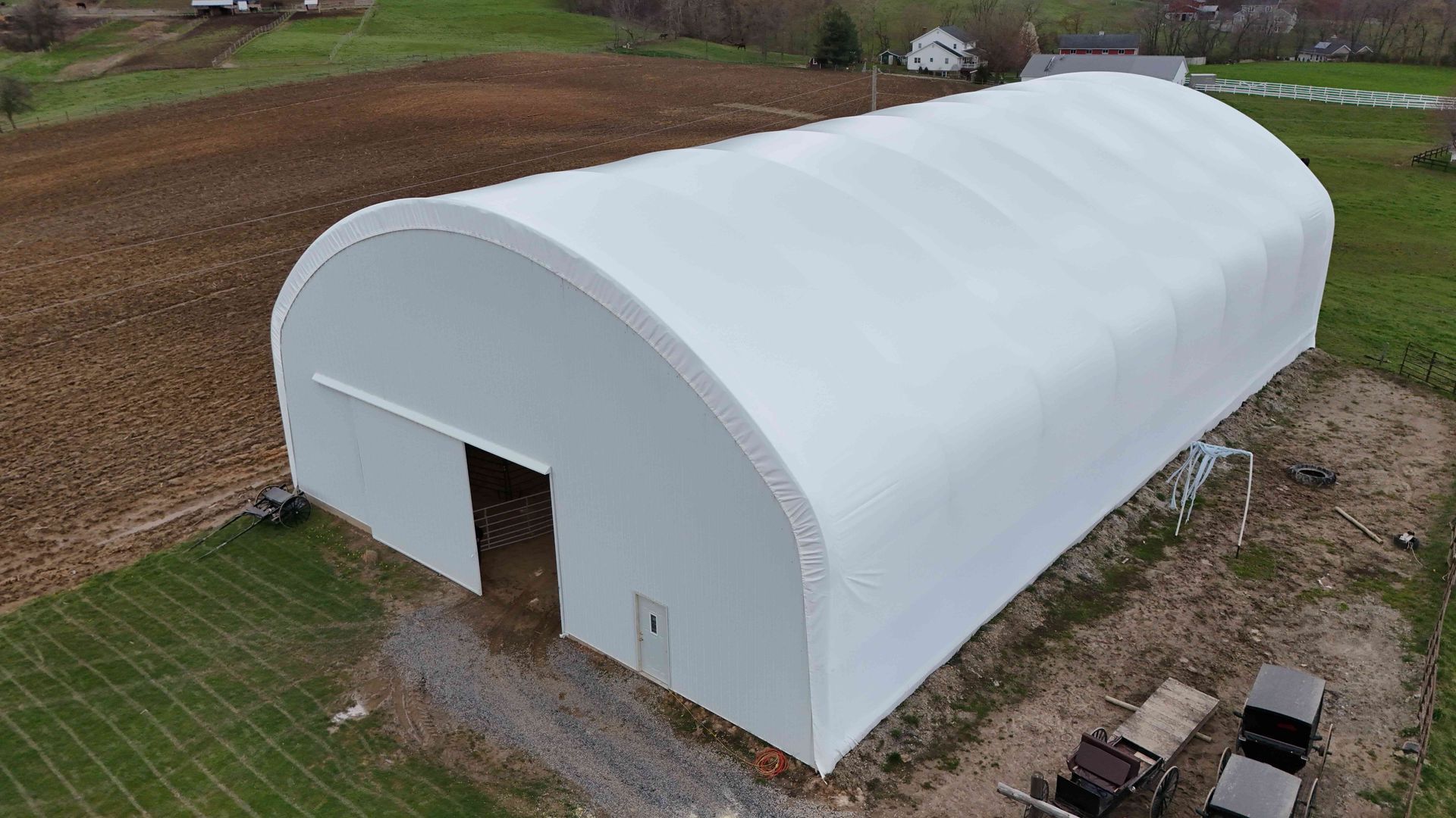 White arched-roof structure with open doorway on a farm, surrounded by fields and equipment.