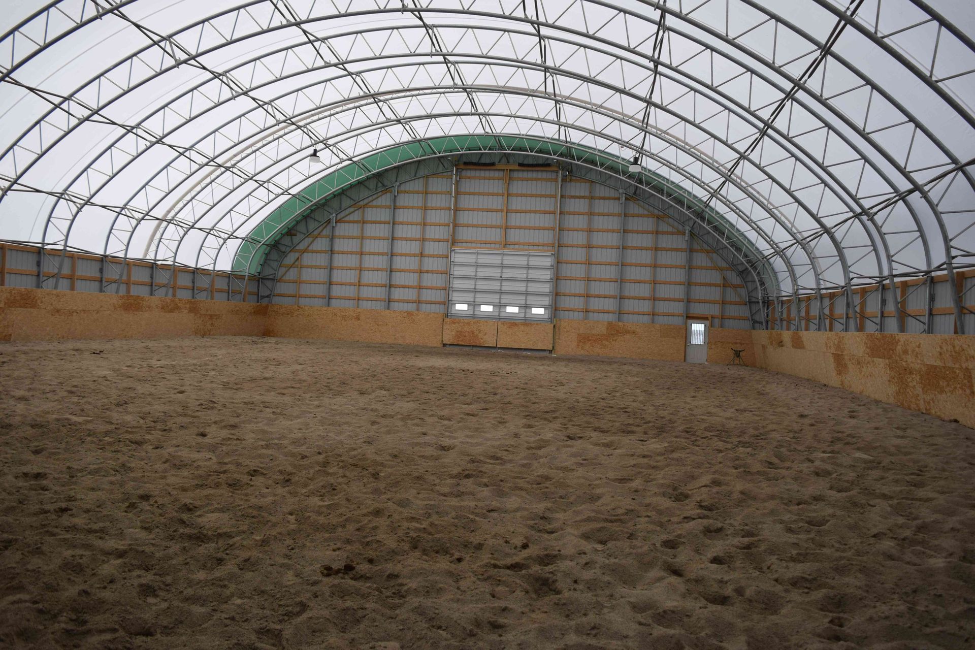 Indoor riding arena with arched metal frame and dirt floor.