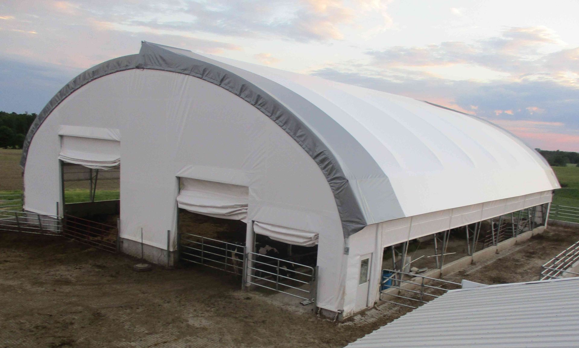 White arched-roof barn with two open doors, a gray trim, and an open side. Evening sky.