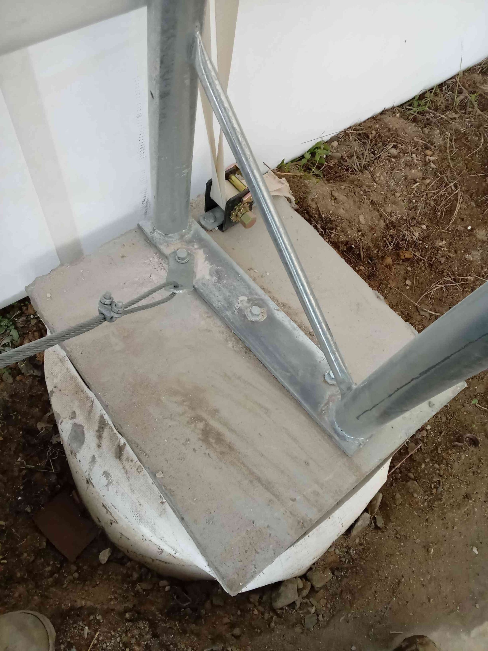 Metal structure base on a concrete slab, near a white wall and dirt ground.