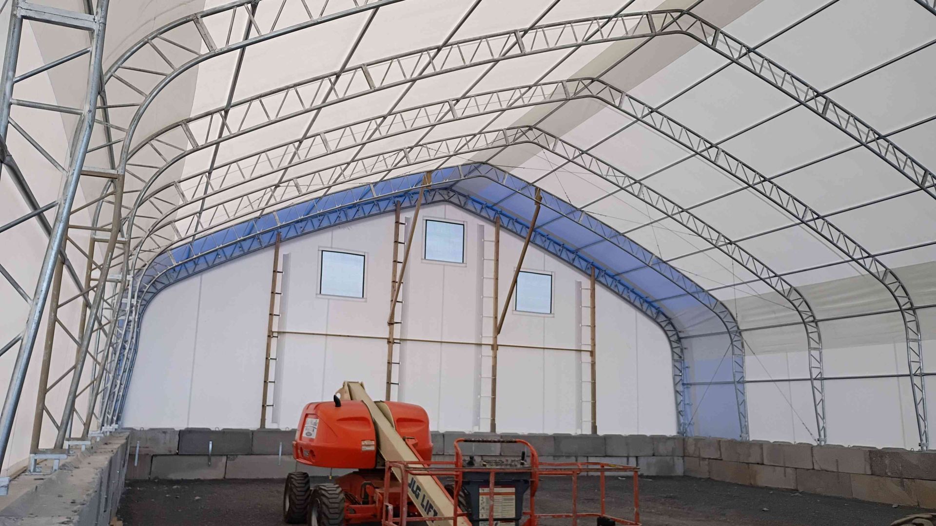 Interior of a large white fabric structure with metal framing, construction equipment, and three small windows.