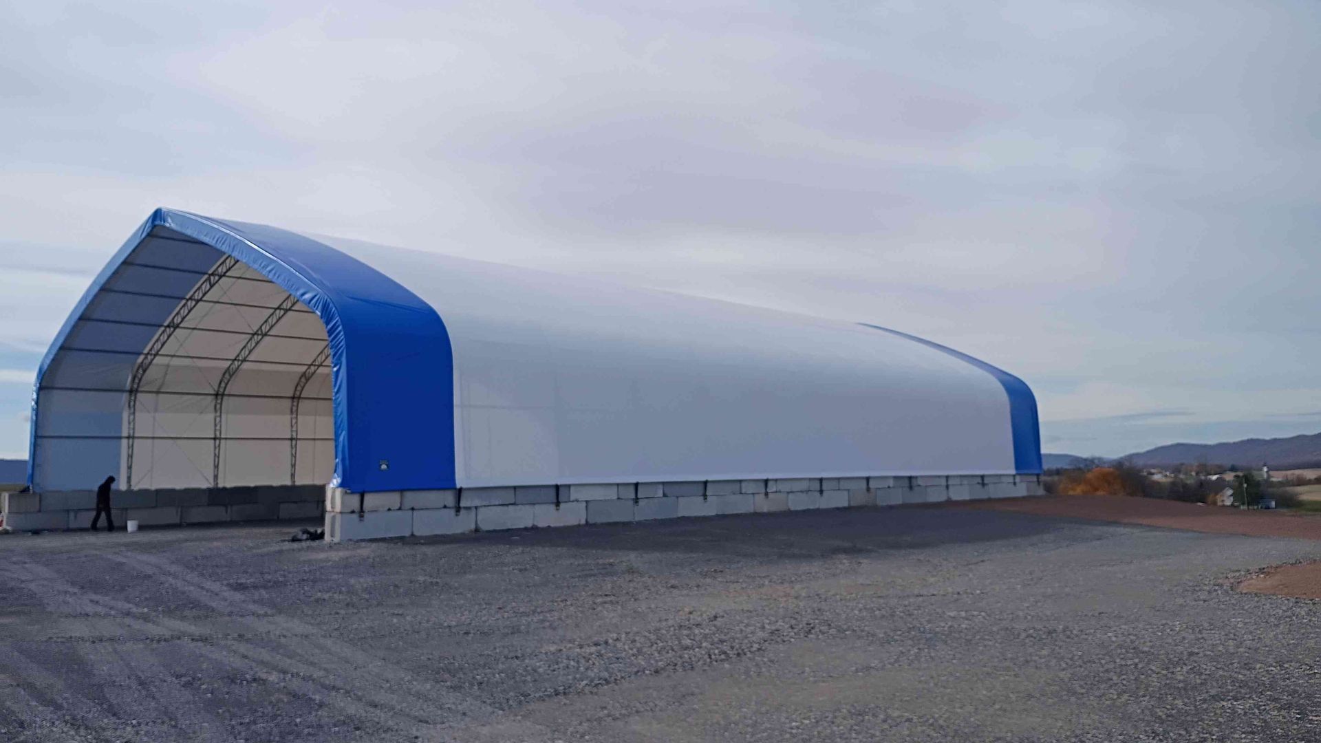 Blue and white arched storage building on a gravel lot; person near the entrance.
