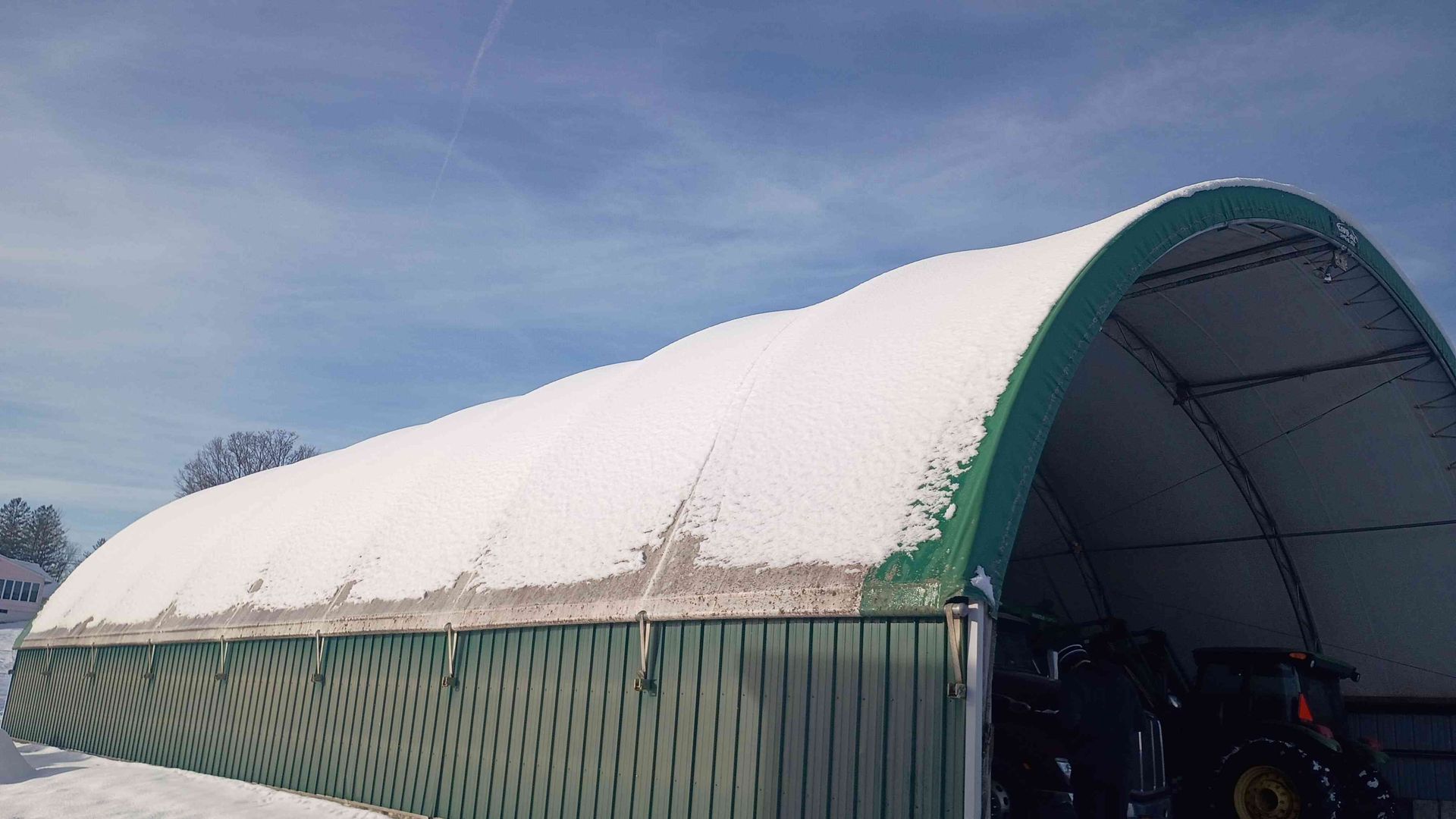 Green arched storage building with snow-covered roof; blue sky.