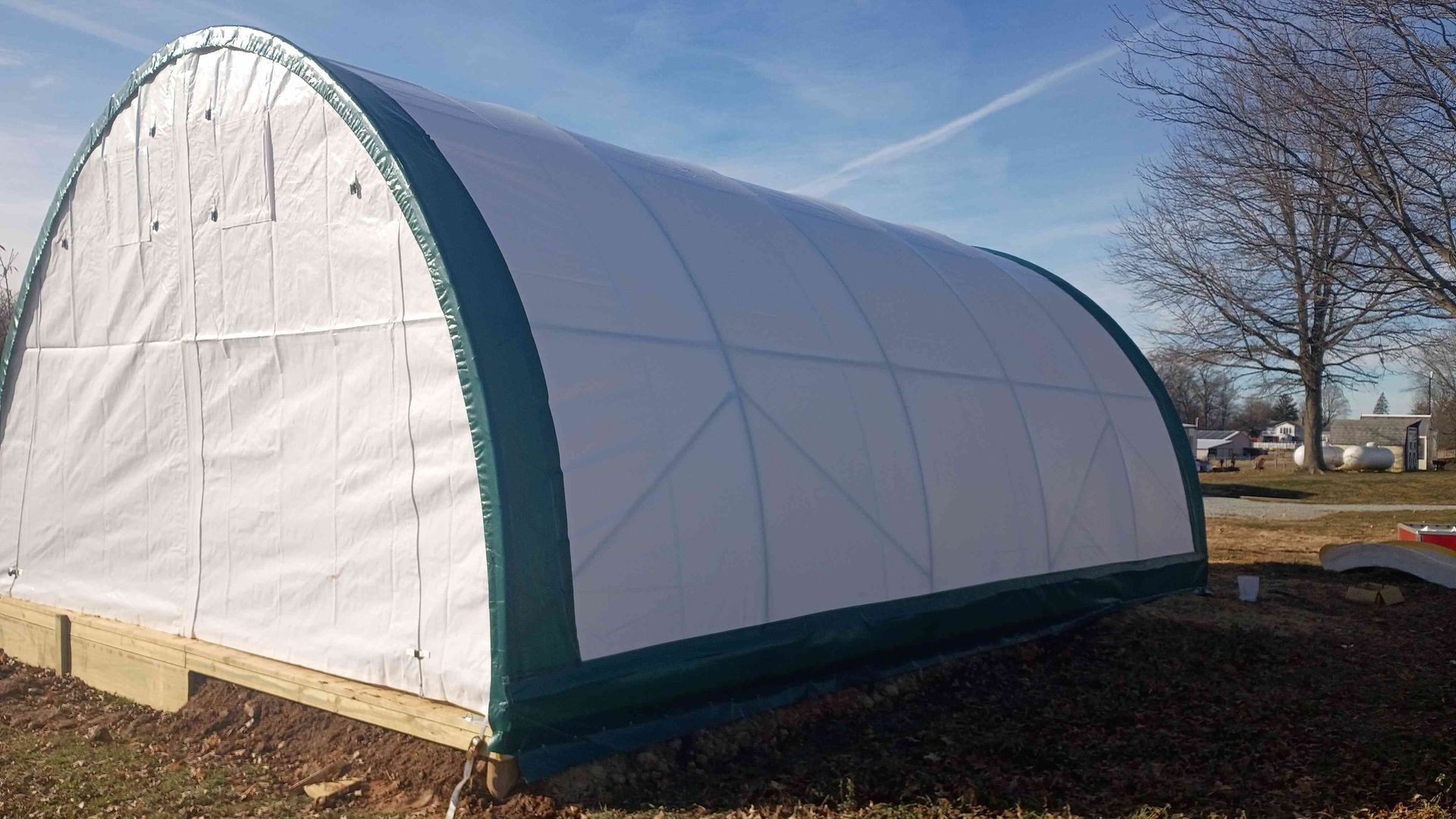 White and green arched greenhouse on a wooden base in a yard with a blue sky.
