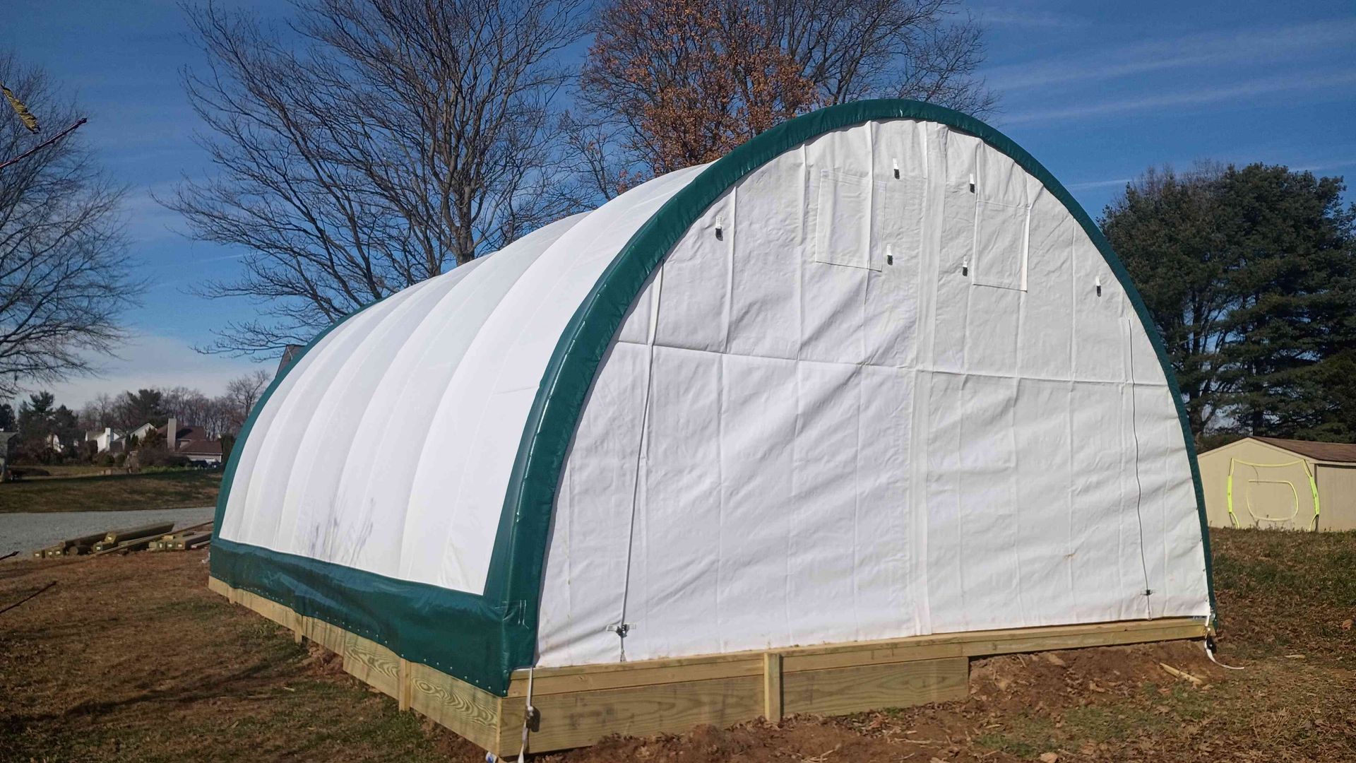 White and green greenhouse structure on a wooden base, outdoors under a blue sky.