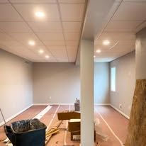 Empty room with exposed ceiling tiles, construction materials, and a large black bin.