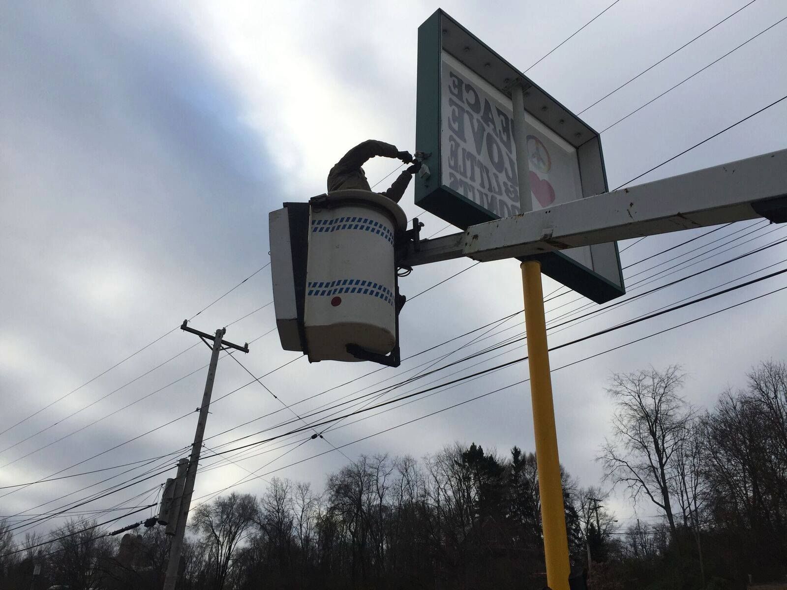 Person in a lift working on a sign. Overcast sky, trees, and power lines in the background.