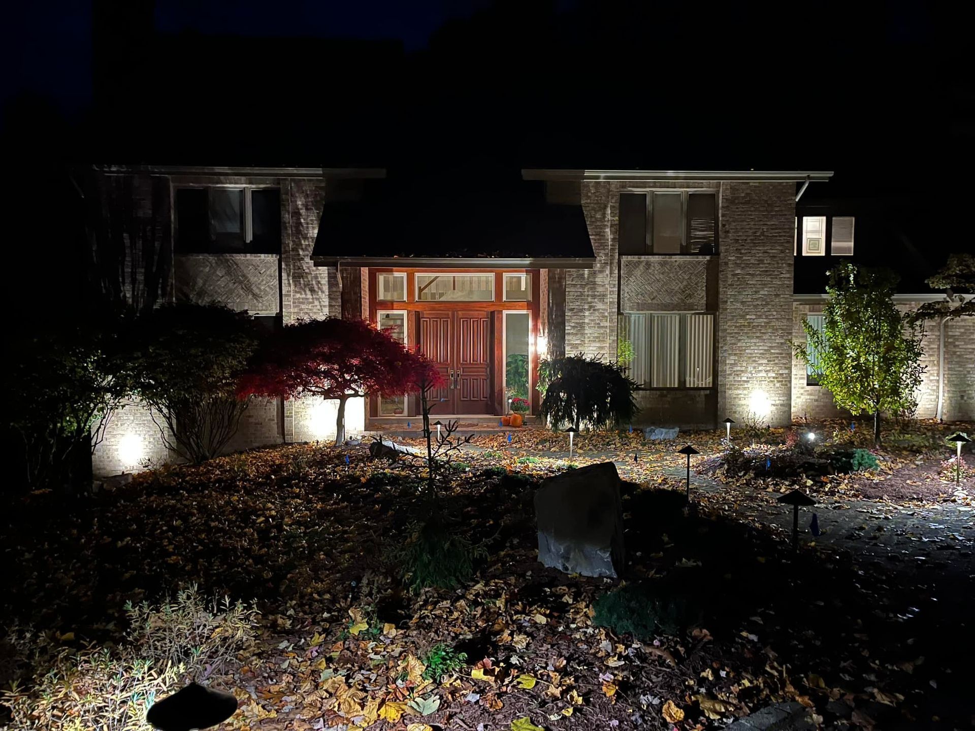A house at night with landscape lighting illuminating the brick facade, door, and front yard foliage.