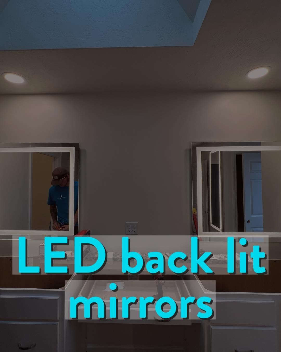 Two mirrors above cabinets with LED backlighting; person reflected in one mirror.