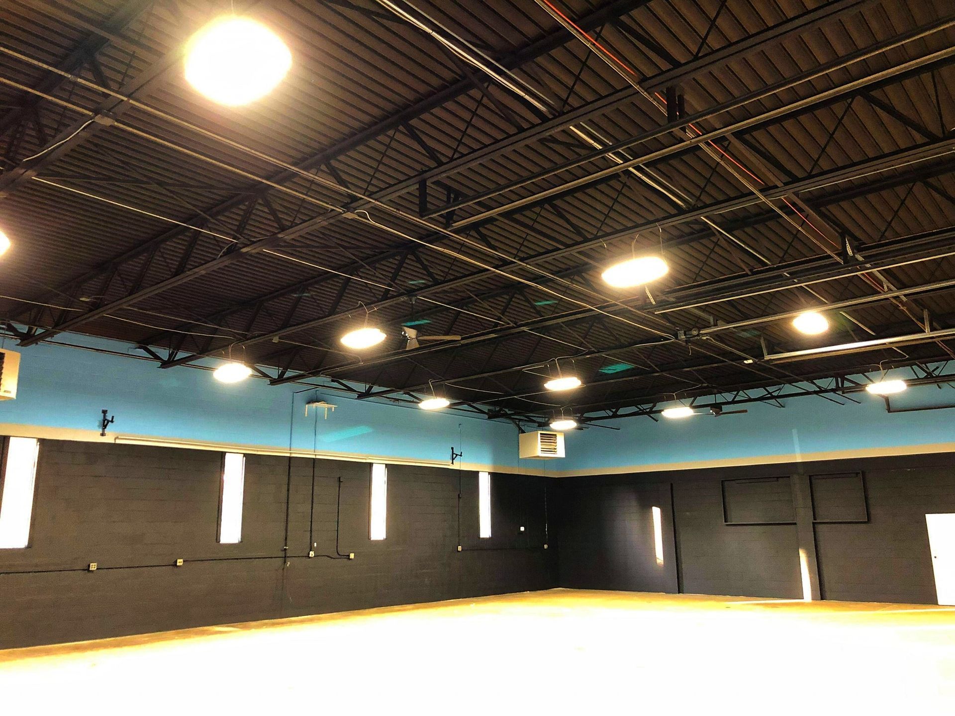 An empty indoor space with a black ceiling, blue wall, and white flooring. Bright overhead lights are visible.