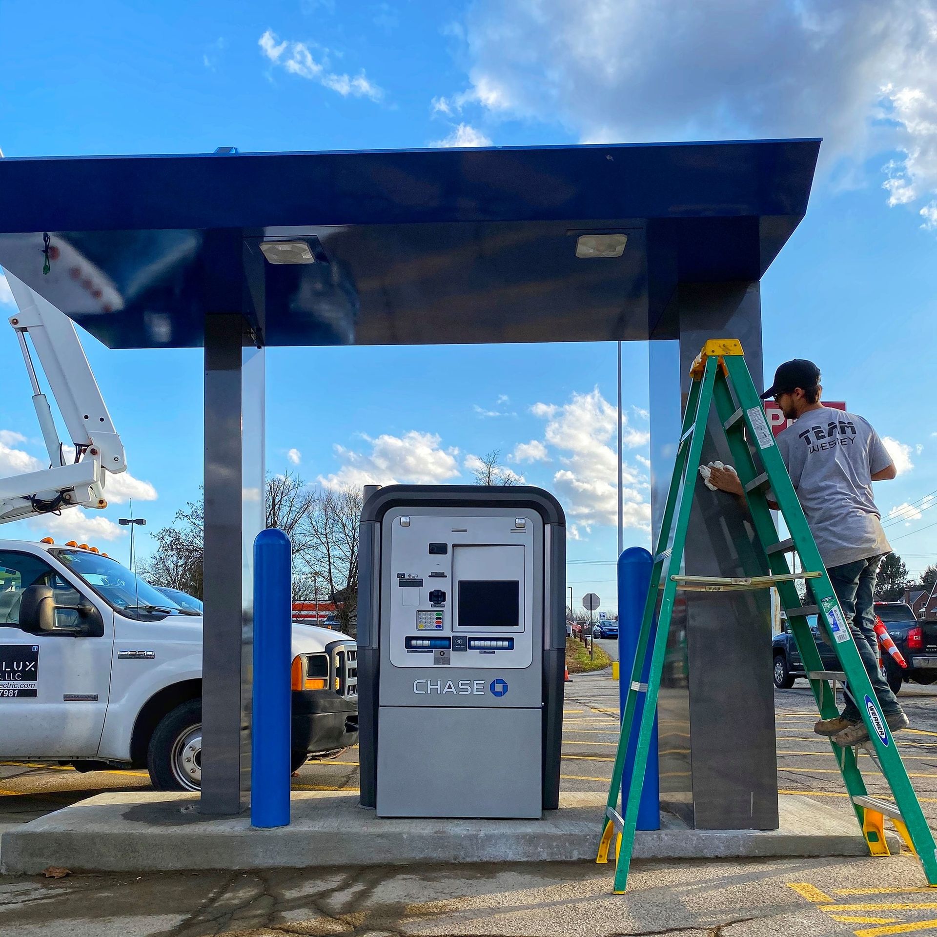 Man on ladder painting ATM canopy. Blue and silver ATM, white truck, blue sky.