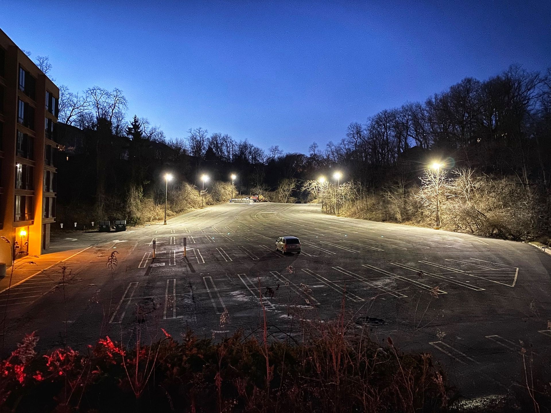 Night view of a parking lot with a single vehicle. Streetlights illuminate the lot and trees in the background.